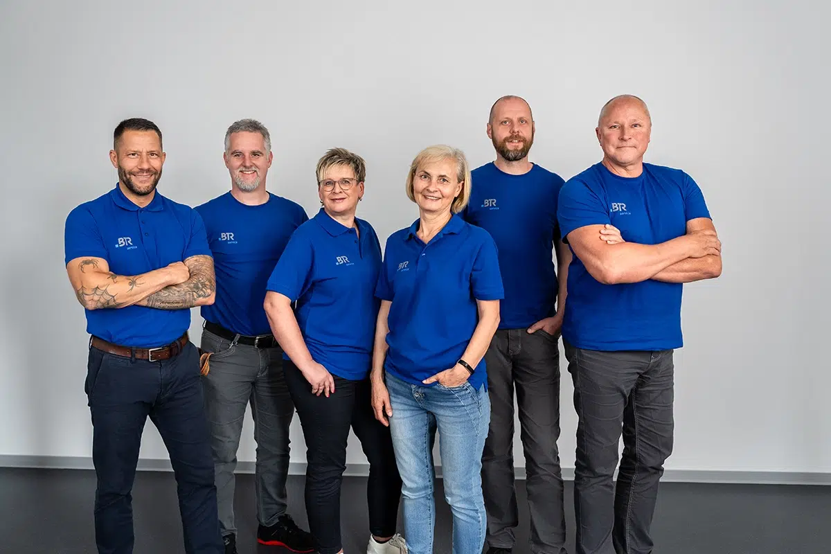 Sechs Teammitglieder in blauen BTR-Servicetrikots posieren lächelnd vor einer grauen Wand.