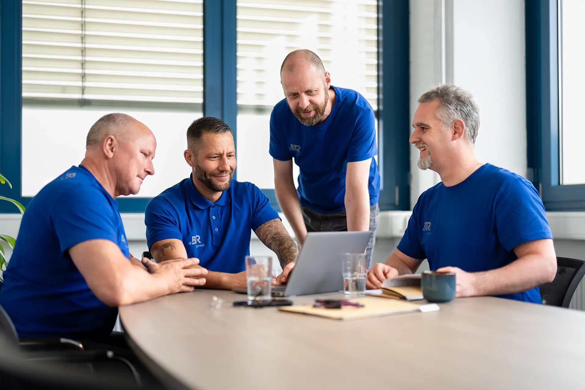 Vier Männer in blauen BTR-Servicetrikots arbeiten gemeinsam lächelnd an einem Laptop in einem Besprechungsraum.