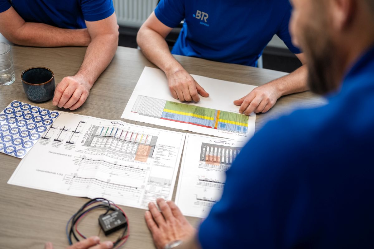 Drei Personen in blauen BTR-Servicetrikots besprechen technische Pläne und Diagramme an einem Tisch.