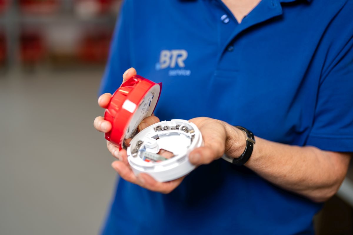 Eine Person in einem blauen BTR-Servicetrikot hält einen geöffneten Rauchmelder, wobei die innere Verkabelung sichtbar ist.