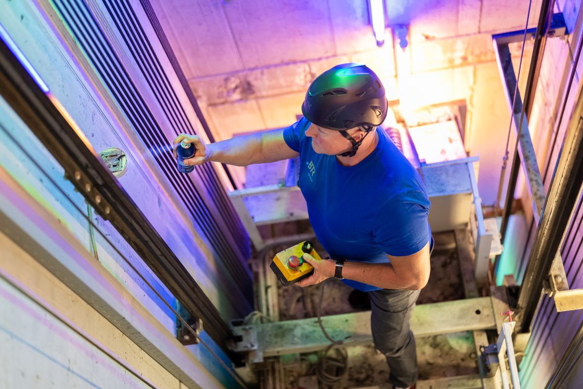 Ein Techniker mit Schutzhelm wartet einen Aufzugsschacht, indem er mit einer Spraydose an den Aufzugsschienen arbeitet und ein Steuergerät in der anderen Hand hält.