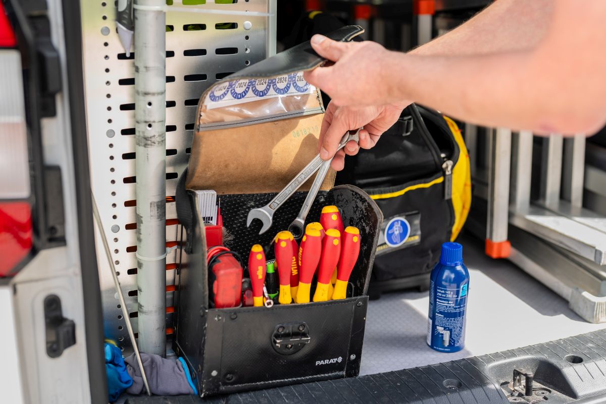 Ein Handwerker greift nach einem Schraubenschlüssel aus einer Werkzeugtasche, die in einem Firmenwagen verstaut ist.