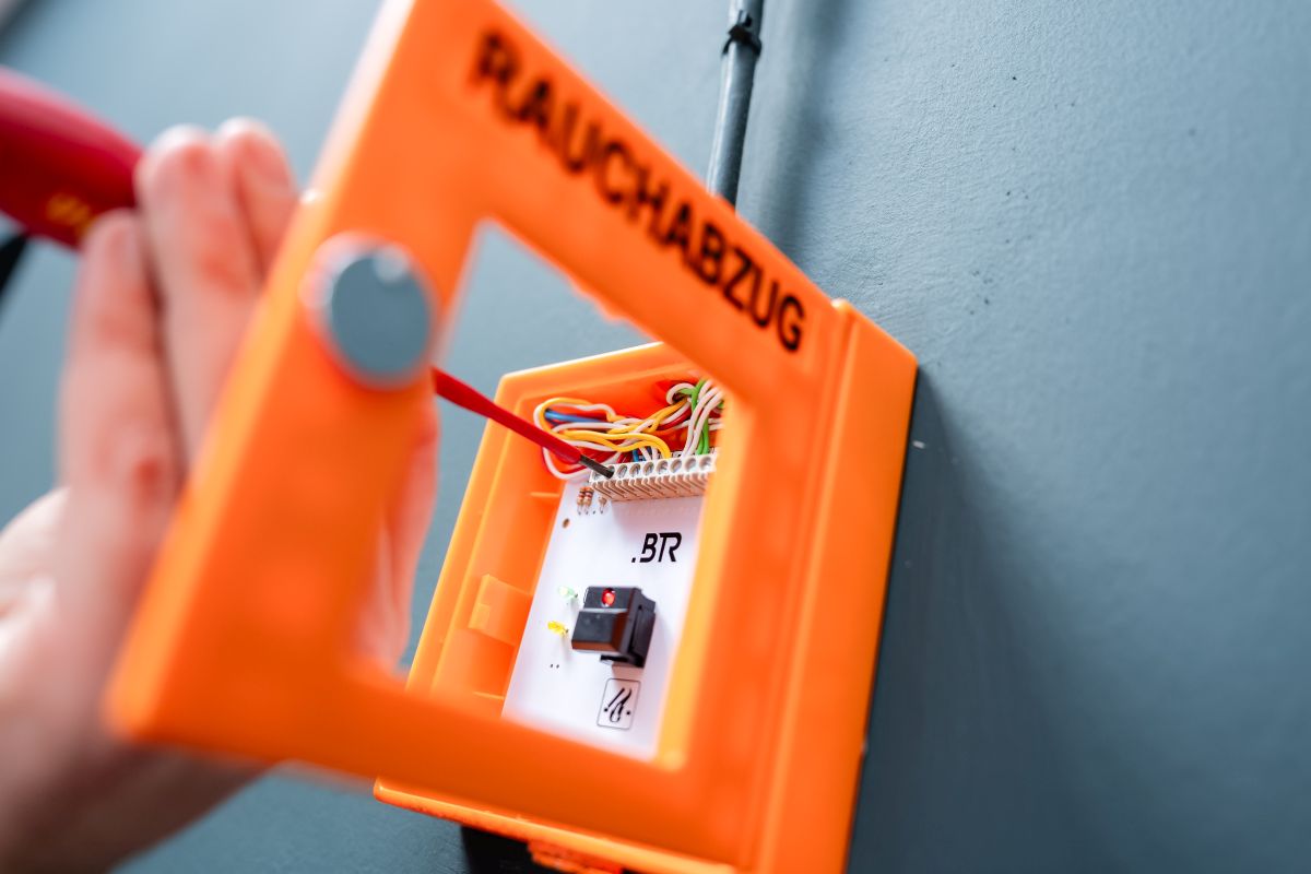 Eine Person installiert oder wartet ein orangefarbenes Rauchabzugssteuergerät an einer Wand und verbindet die Drähte im Inneren.