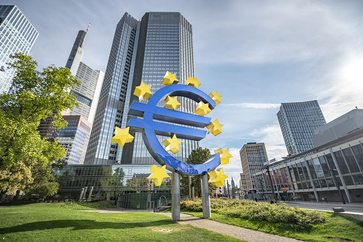 Große Euro-Skulptur mit gelben Sternen im Vordergrund, umgeben von modernen Hochhäusern in einem Geschäftsviertel bei sonnigem Wetter.