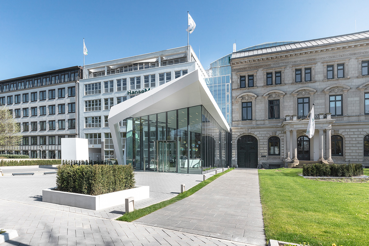 Moderner Glasbau neben historischen Gebäuden mit gepflegter Grünfläche und blauem Himmel im Hintergrund.