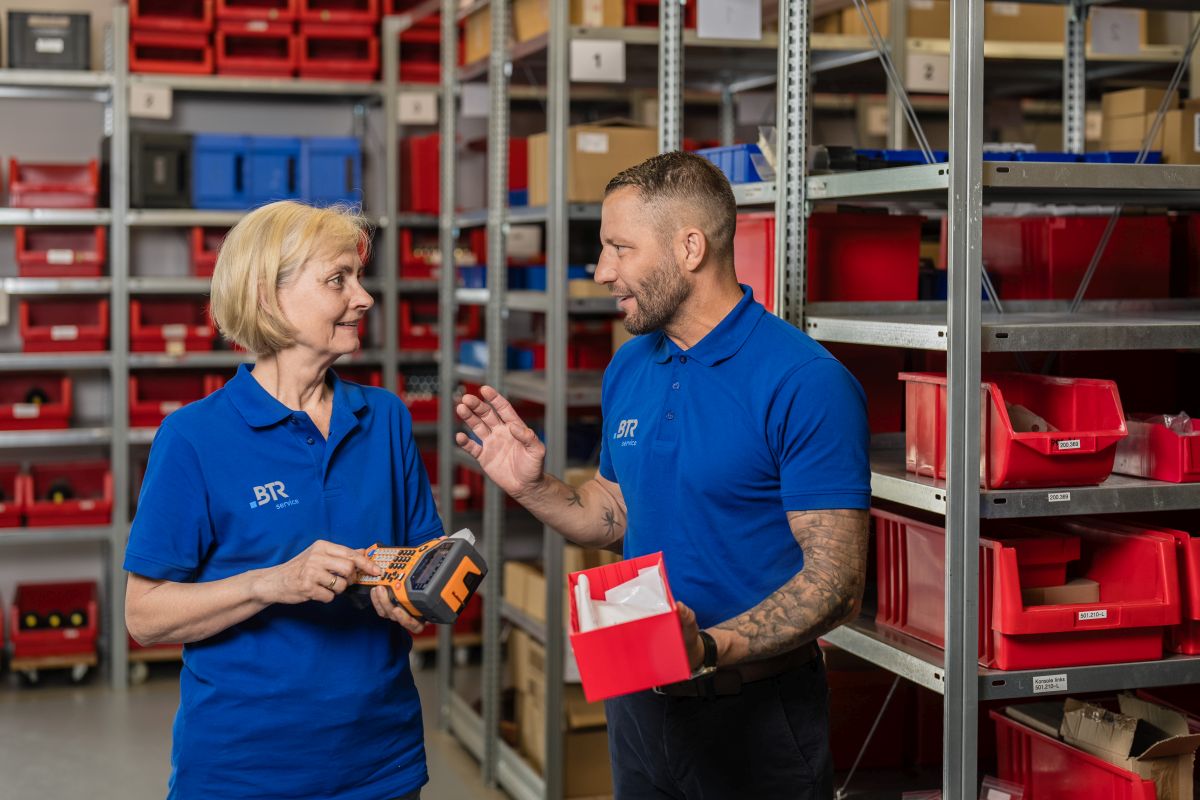 Zwei Mitarbeitende in blauen BTR-Servicetrikots unterhalten sich in einem Lager mit Regalen voller roter Kisten. Einer hält ein Beschriftungsgerät, der andere eine rote Kiste mit kleinen Teilen.