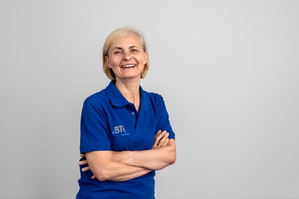 Lächelnde Frau in einem blauen BTR-Servicetrikot mit verschränkten Armen vor einer grauen Wand.