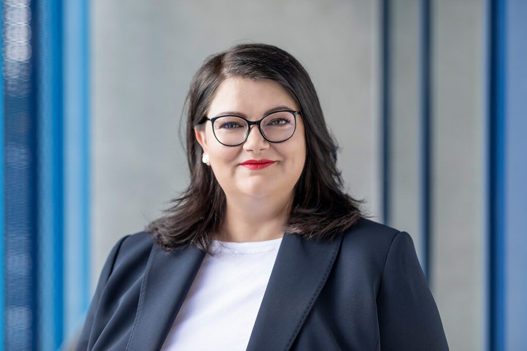 Porträt einer Frau mit dunklen Haaren, Brille und rotem Lippenstift, die in einem dunkelblauen Blazer vor einem verschwommenen Hintergrund lächelt.